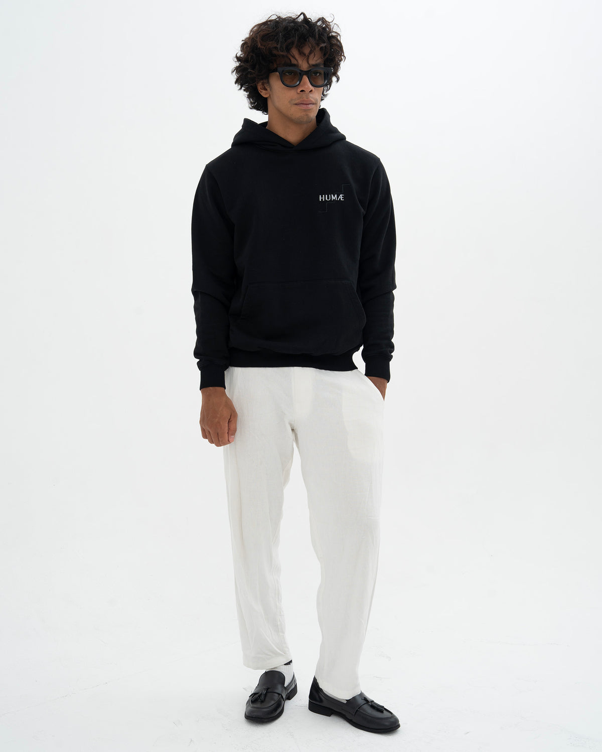 Man wearing the HUMÆ Founder Hoodie in Deep Black with white trousers, full-body front view