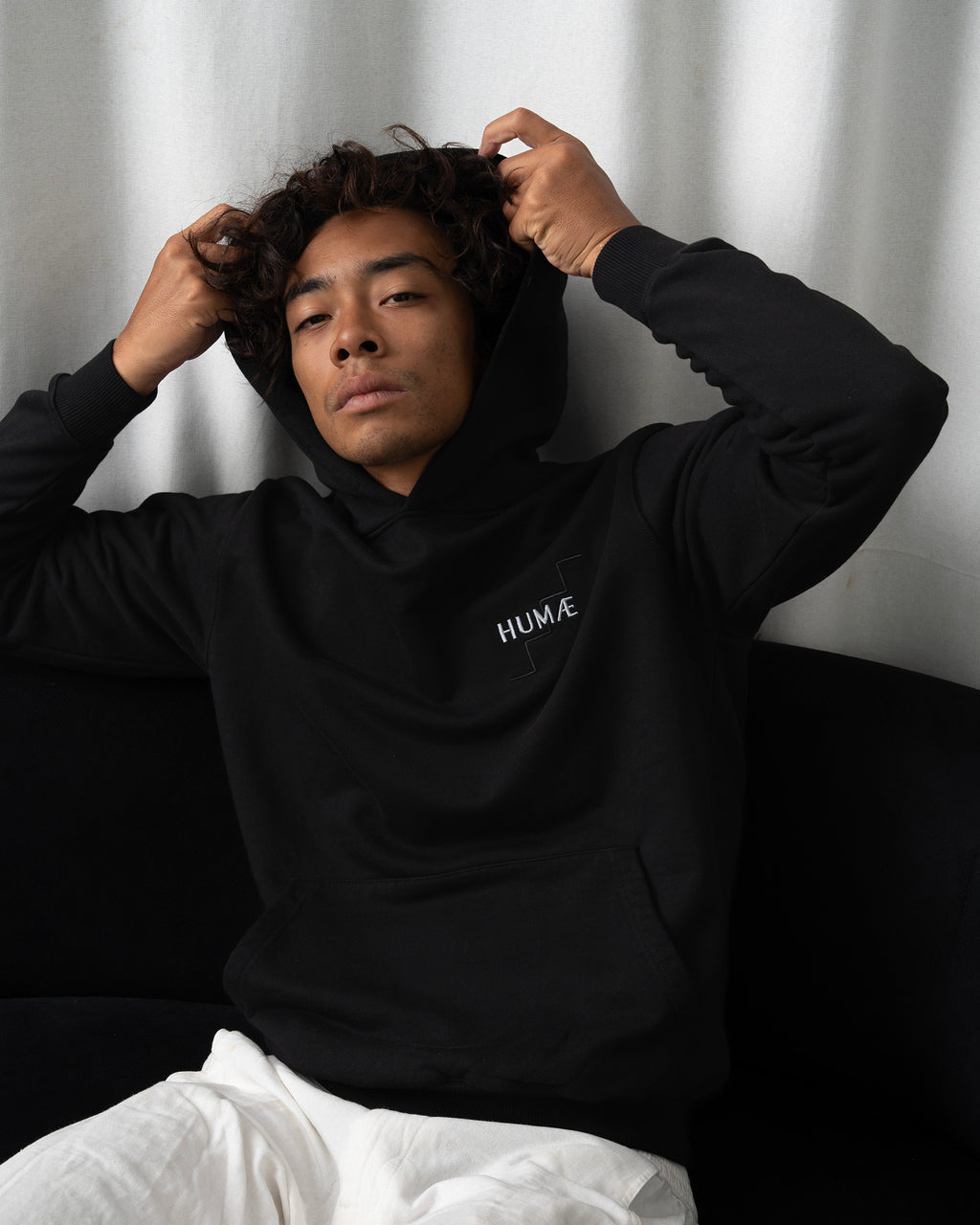 Man wearing the HUMÆ Founder Hoodie in Deep Black, seated while adjusting the hood