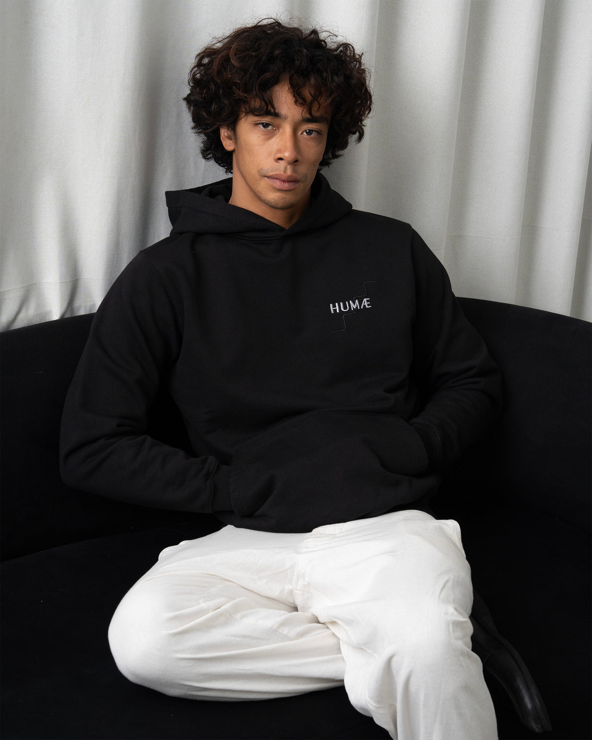 Man wearing the HUMÆ Founder Hoodie in Deep Black, seated in a relaxed pose.