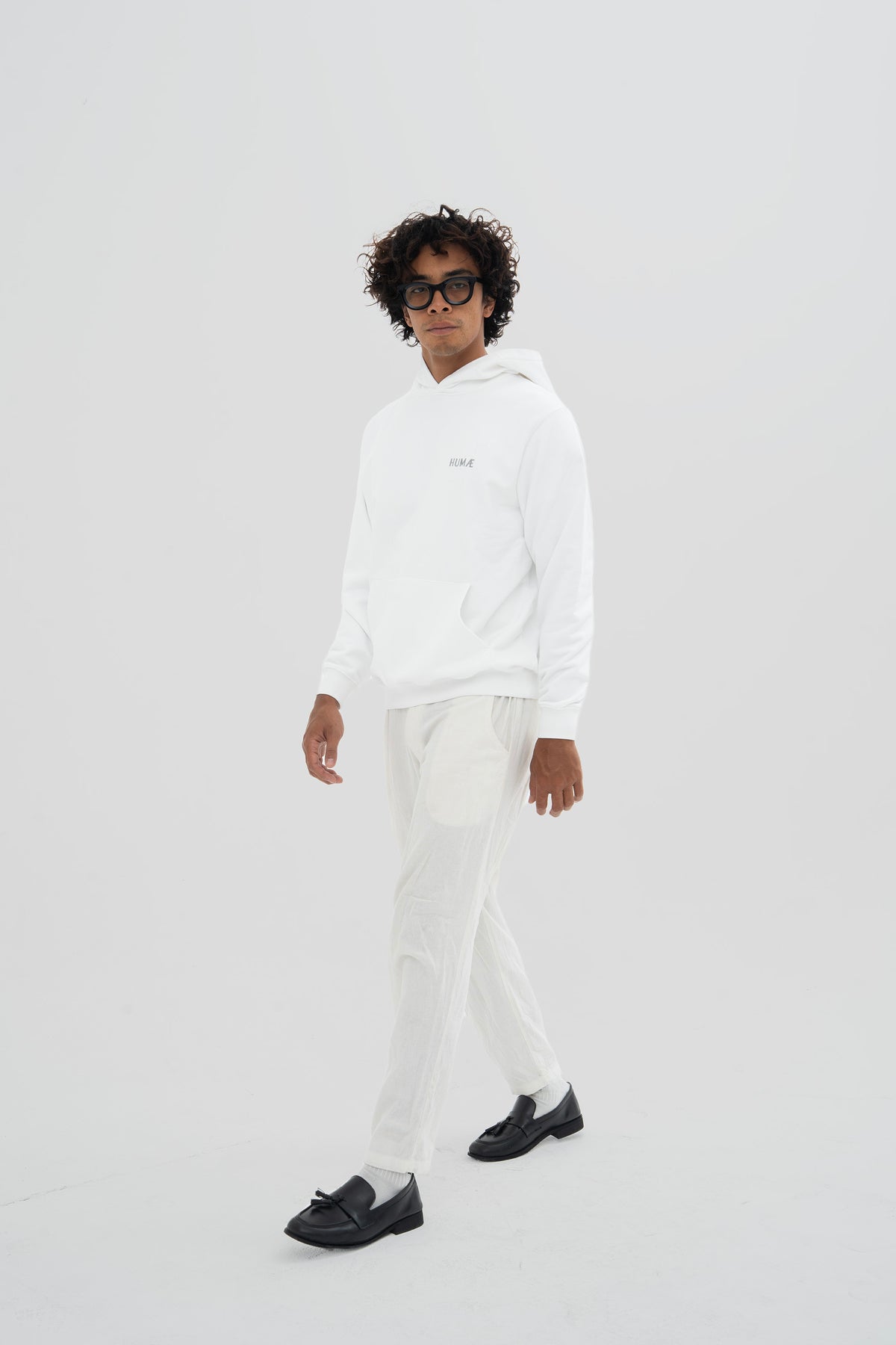 Man wearing the HUMÆ Founder Hoodie in Optical White with white trousers, shown walking in a full-body view