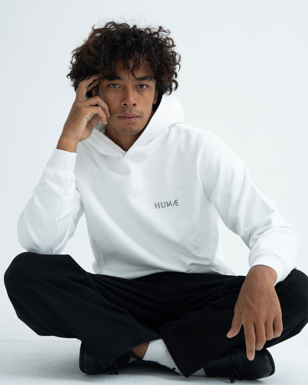 Man wearing the HUMÆ Founder Hoodie in Optical White, seated cross-legged in a relaxed pose
