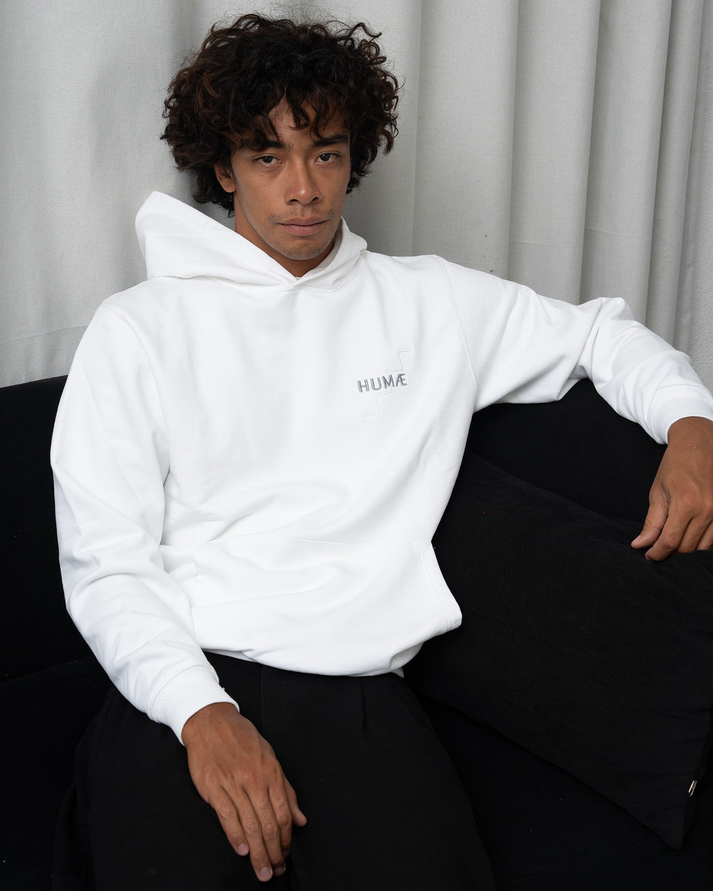 Man wearing the HUMÆ Founder Hoodie in Optical White, seated on a black sofa in a relaxed posE