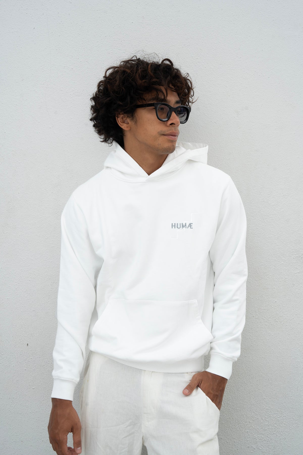 Man wearing the HUMÆ Founder Hoodie in Optical White with white trousers, standing and looking to the side