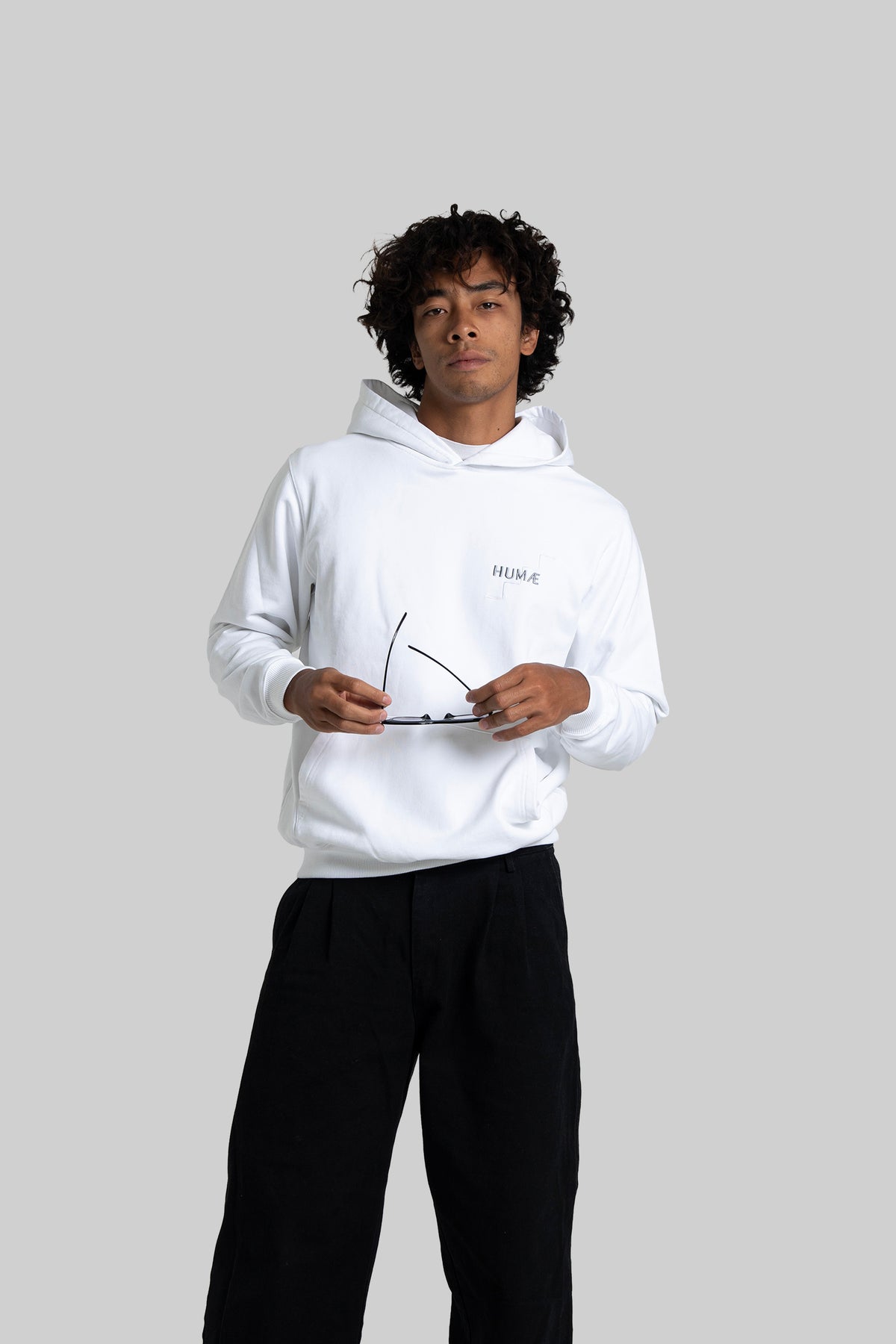 Man wearing the HUMÆ Founder Hoodie in Optical White, standing while holding sunglasses